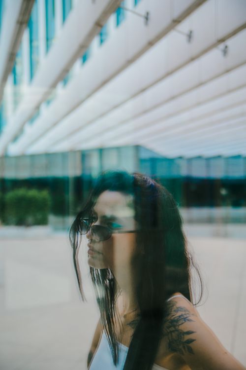 Free A thoughtful portrait of a woman with sunglasses and tattoo reflecting on glass in a modern urban context. Stock Photo