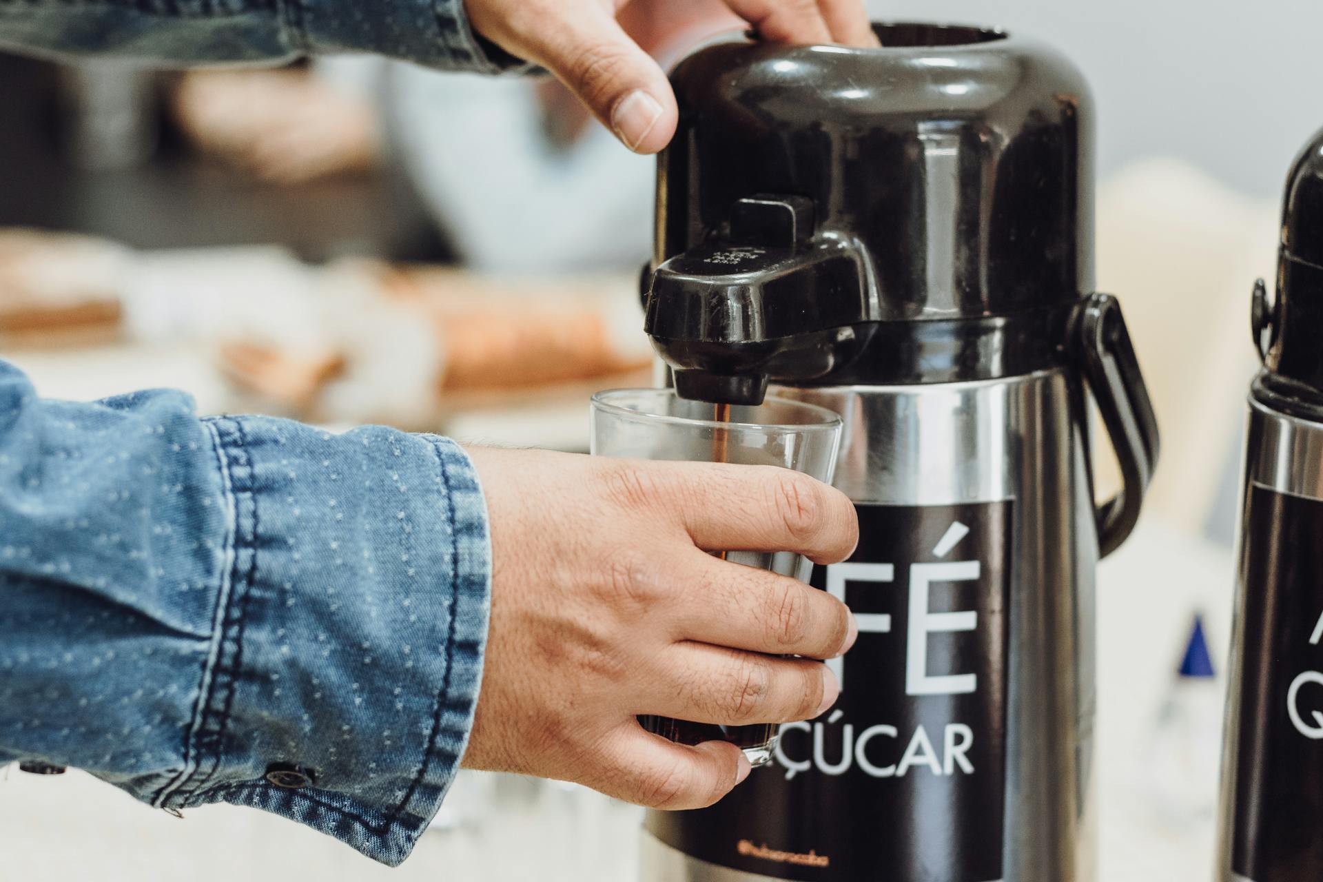 Person pouring coffee from a stainless steel dispenser into a glass.