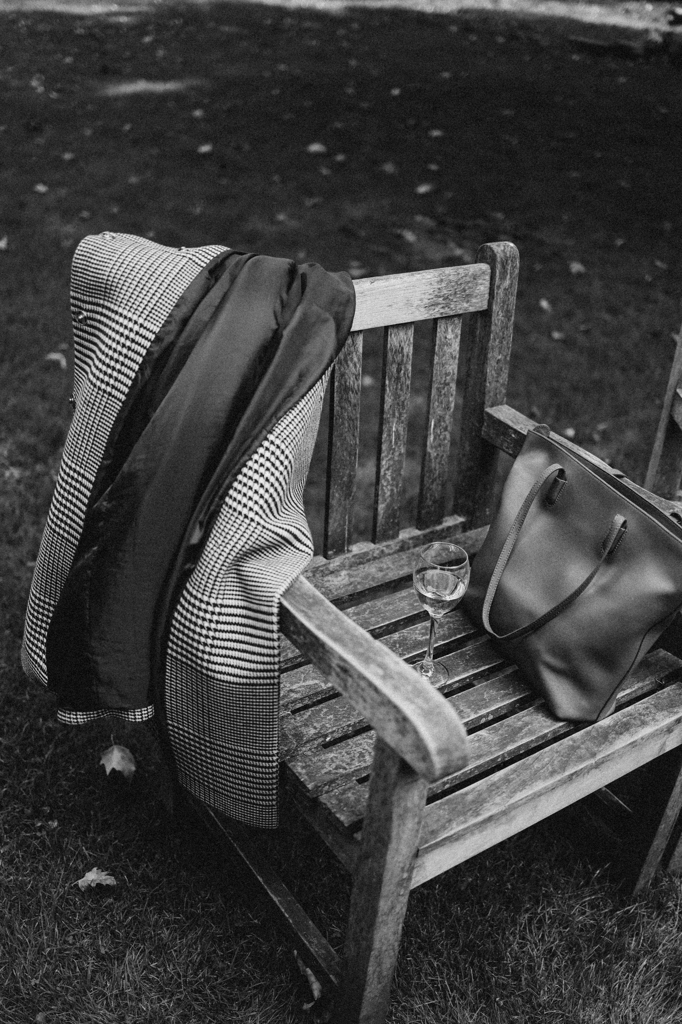 A black and white shot of a chair with a coat, handbag, and wine glass outdoors.
