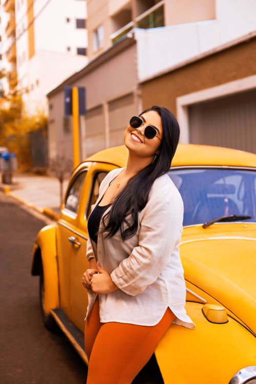 Free Woman with sunglasses leans against a classic yellow Volkswagen Beetle on an urban street in Bauru, Brazil. Stock Photo