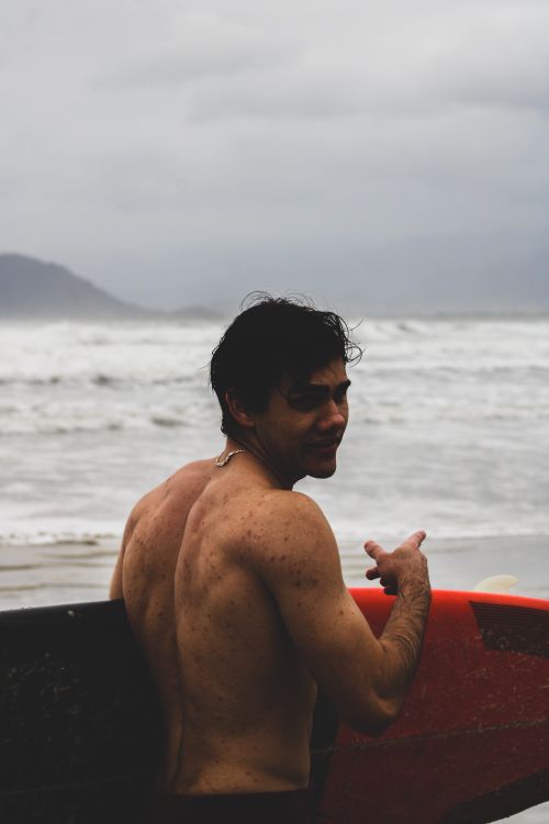 Free Shirtless surfer with a red surfboard on a cloudy day at the beach, looking out to the ocean. Stock Photo