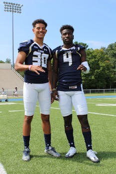 Two athletes in football uniforms pose on a sunny outdoor field.