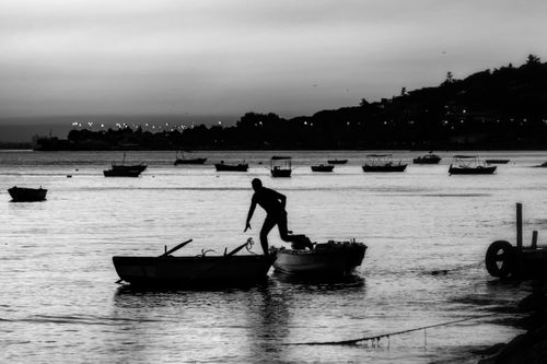 Free A person standing between boats on a serene shoreline at dusk in monochrome tones. Stock Photo