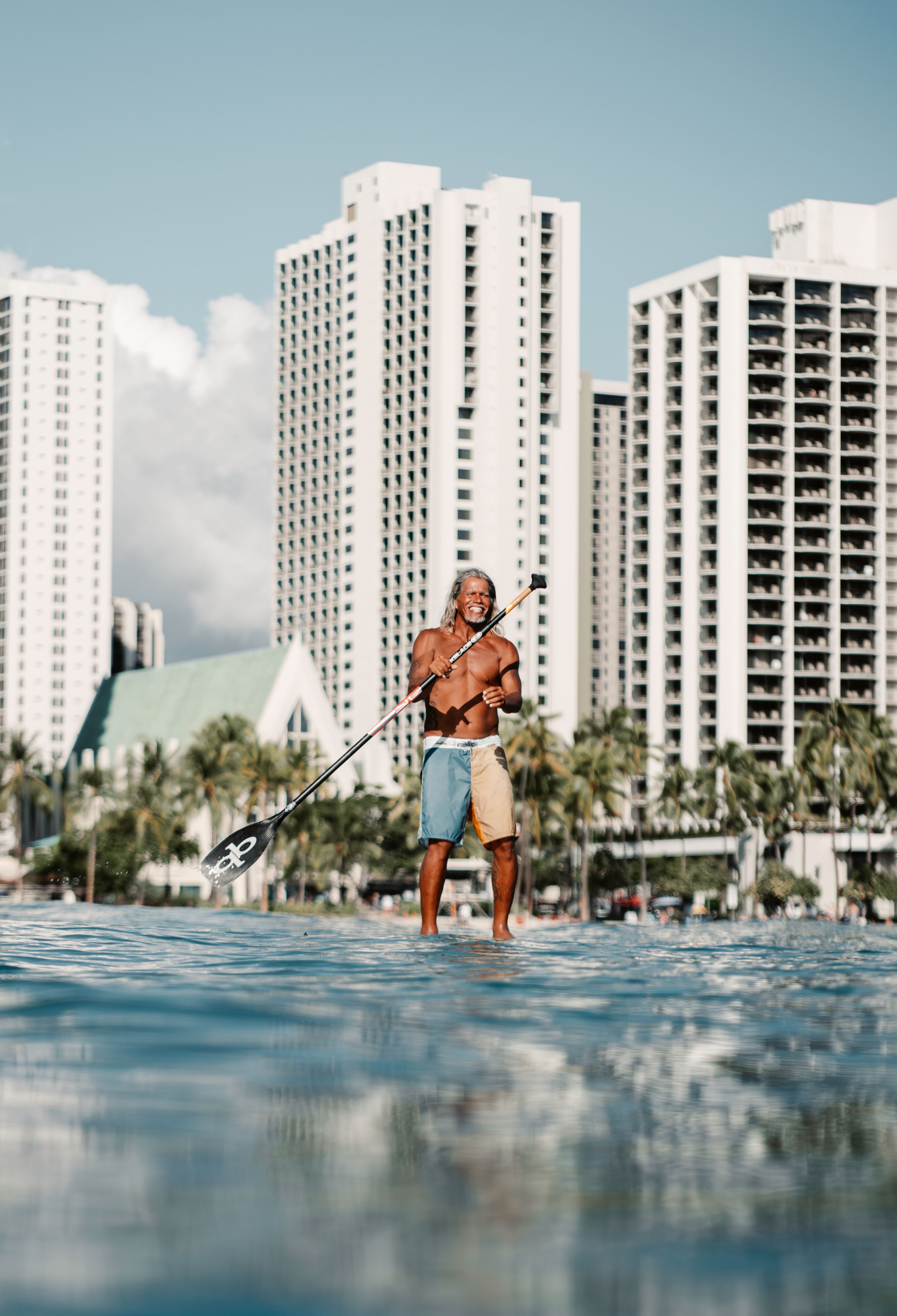 A shirtless man paddleboarding in the ocean near tall urban buildings on a sunny day.