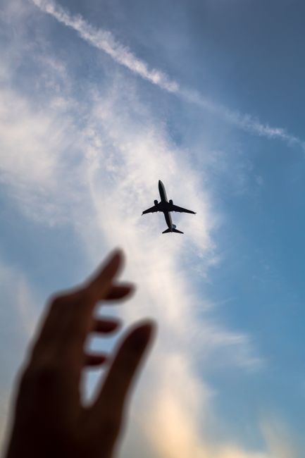 Silhouette of a hand reaching towards a flying airplane against a cloudy sky.