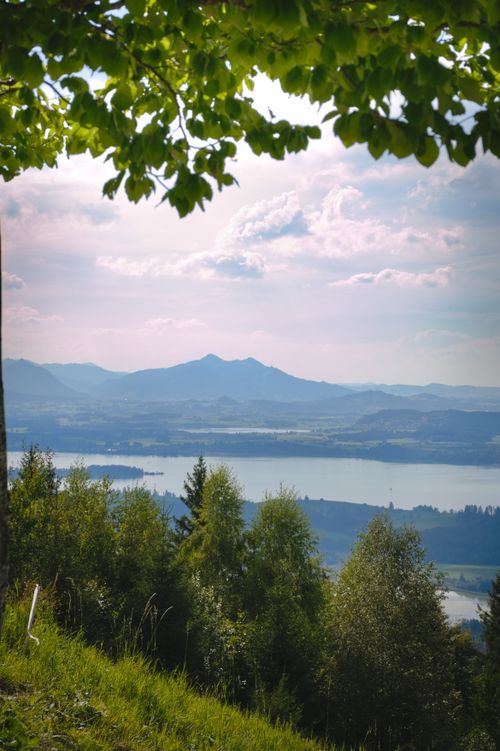 Free Scenic view of Buchenberg with lush greenery, serene lake, and distant mountains under a cloudy sky. Stock Photo