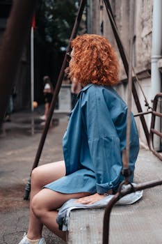 Woman with curly hair in blue oversized shirt sitting on a metal bench in an urban setting.