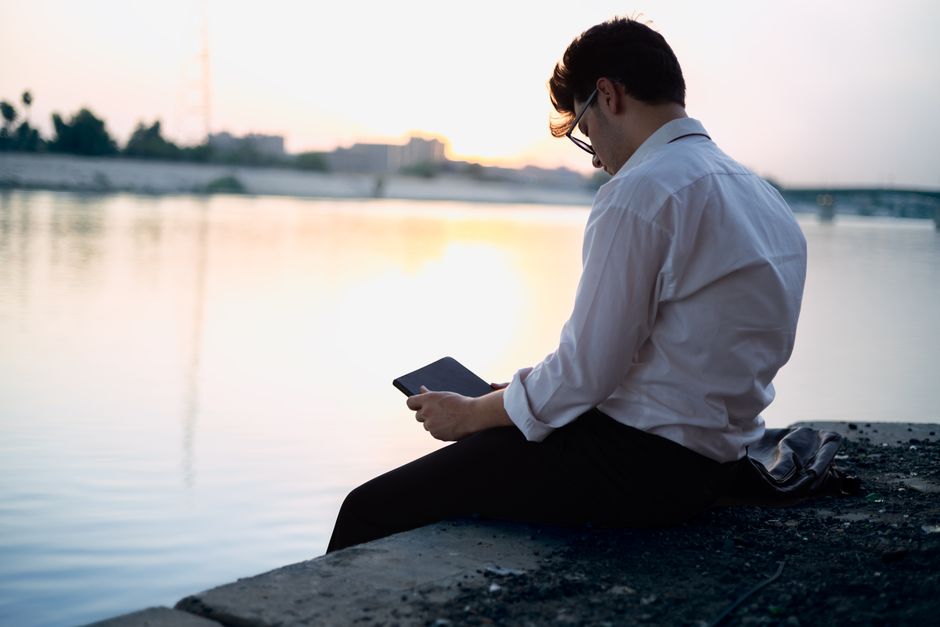 A businessman seated by a riverbank, reading on a tablet as the sun sets, creating a peaceful and reflective scene.