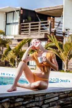 Woman in swimwear enjoying a sunny day by the pool, holding a cup of coffee.