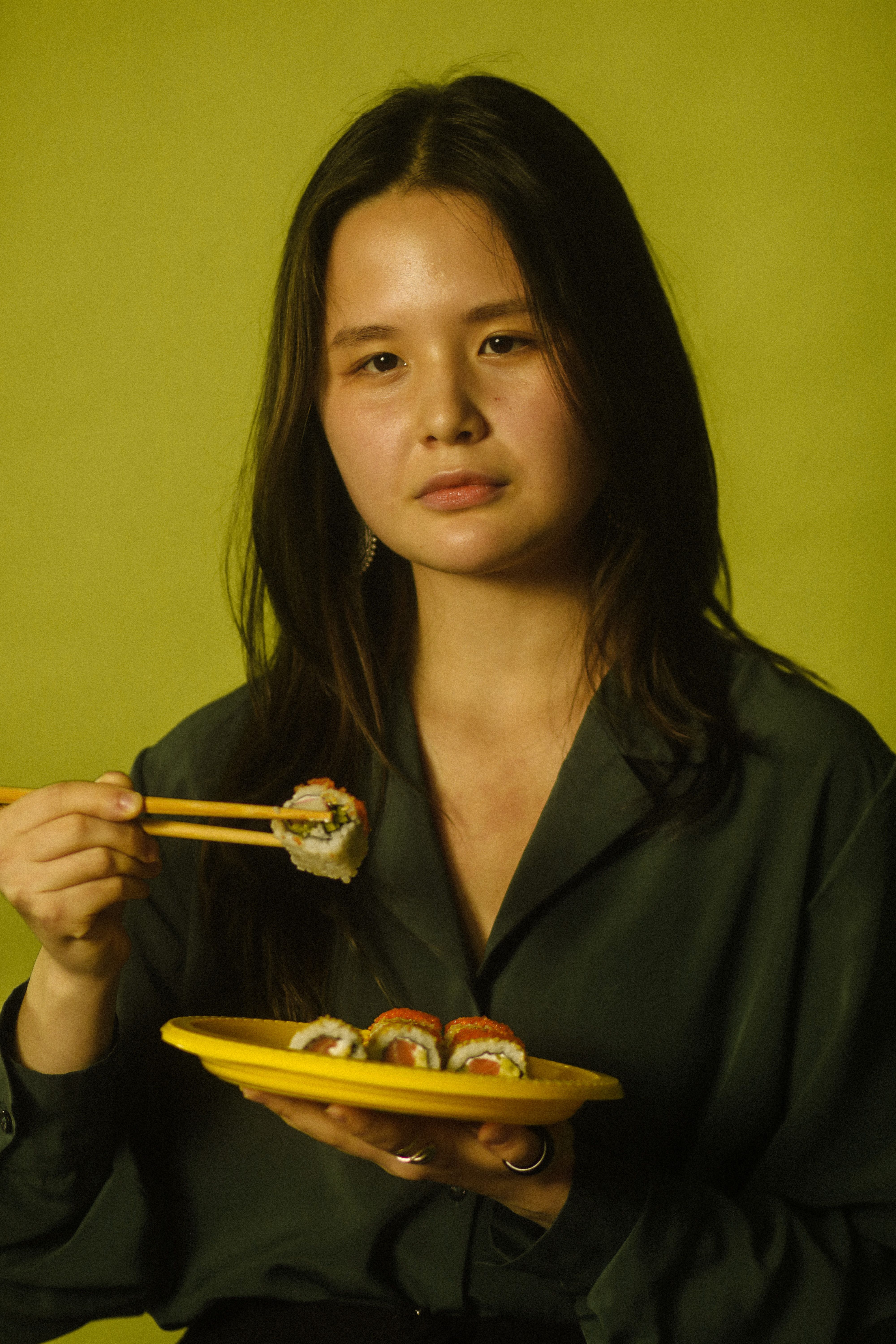 Asian woman holding a sushi plate with chopsticks in front of a green background.