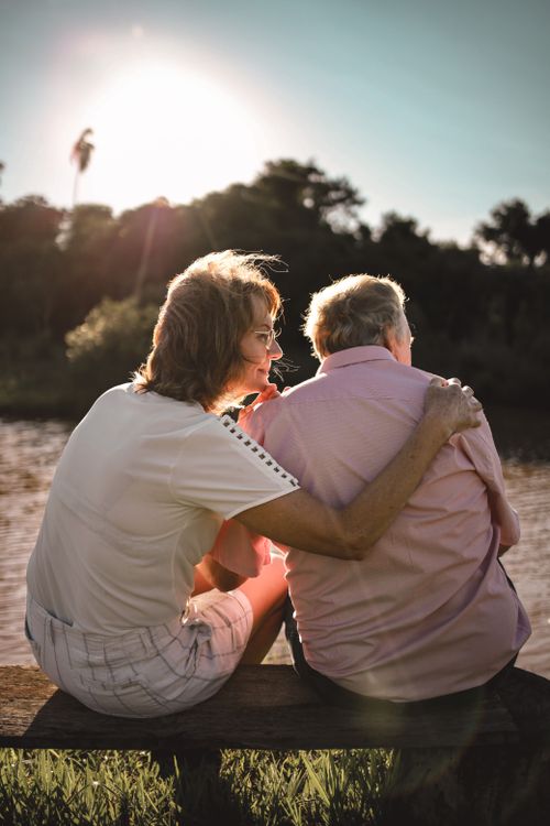 Free A heartwarming scene of a senior couple embracing on a riverside bench during sunset. Stock Photo