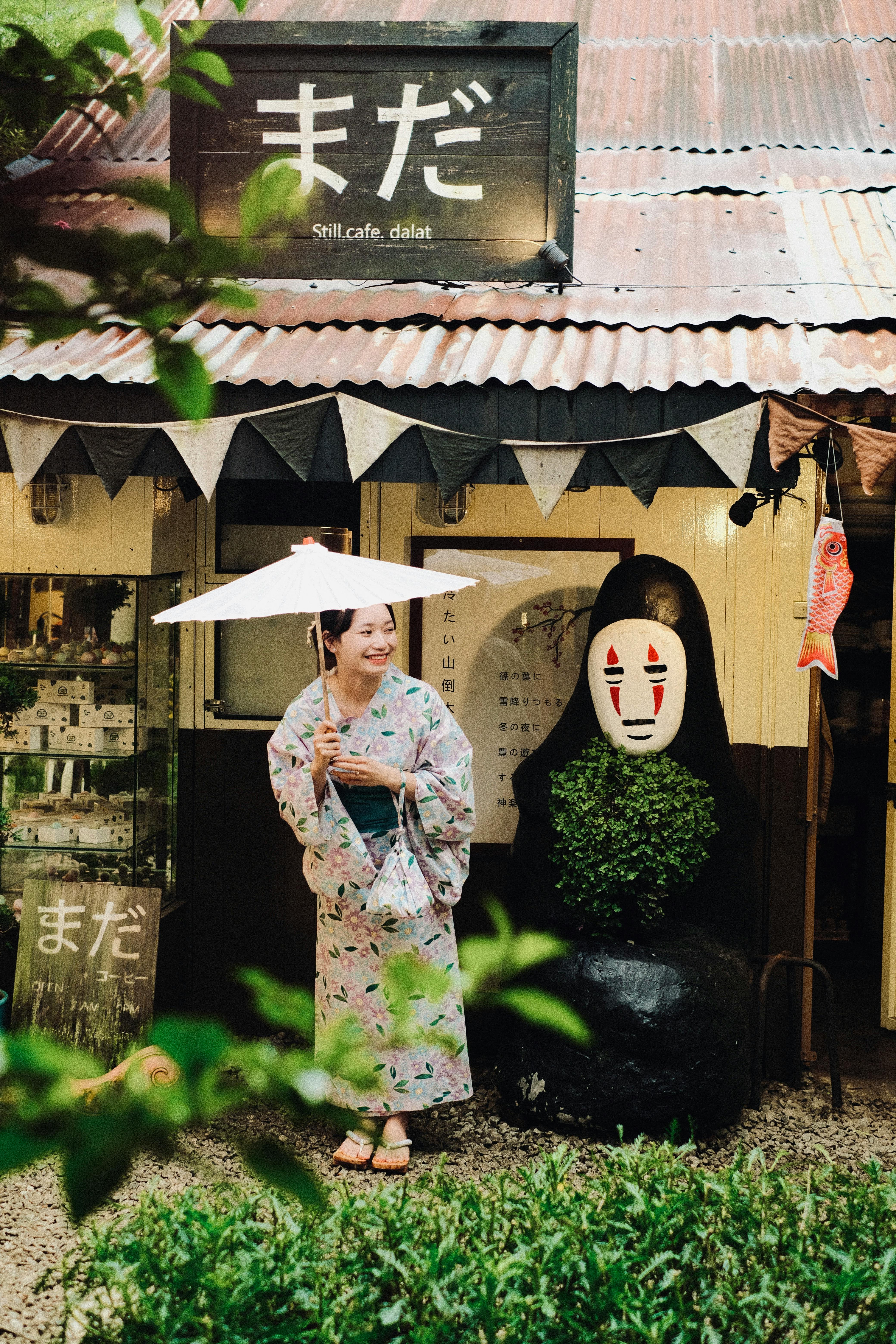 Asian woman in traditional attire smiling under an umbrella at a charming Japanese cafe.