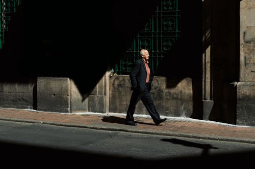 Free A well-dressed man walks in a shaded street in Bogotá, Colombia, conveying urban elegance. Stock Photo