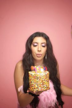Woman celebrating birthday, blowing candles on a colorful cake against a pink background.
