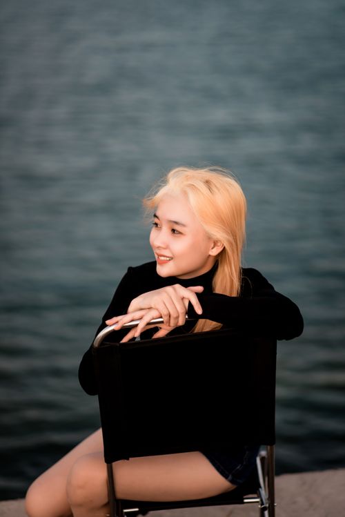 Free Portrait of a smiling blonde woman in black, seated near serene water outdoors. Stock Photo