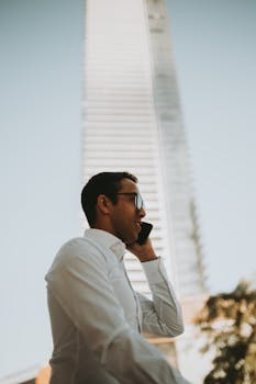 Adult businessman in white shirt making a phone call beside a skyscraper. Low angle shot outdoors.