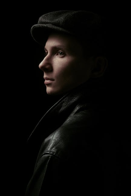 Side profile portrait of man in leather jacket and hat against black background.