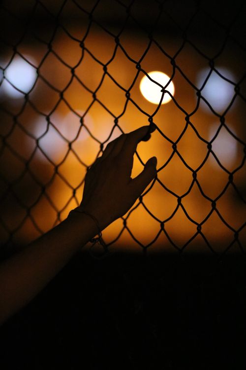 Free A close-up of a hand touching a chain link fence with blurred lights in the background at night. Stock Photo
