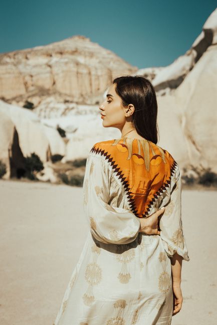 A woman in a traditional dress stands side view under a clear blue sky against rock mountains.