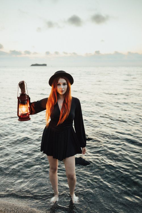 Free A woman in a black dress and hat holds a lantern standing on a twilight beach. Stock Photo