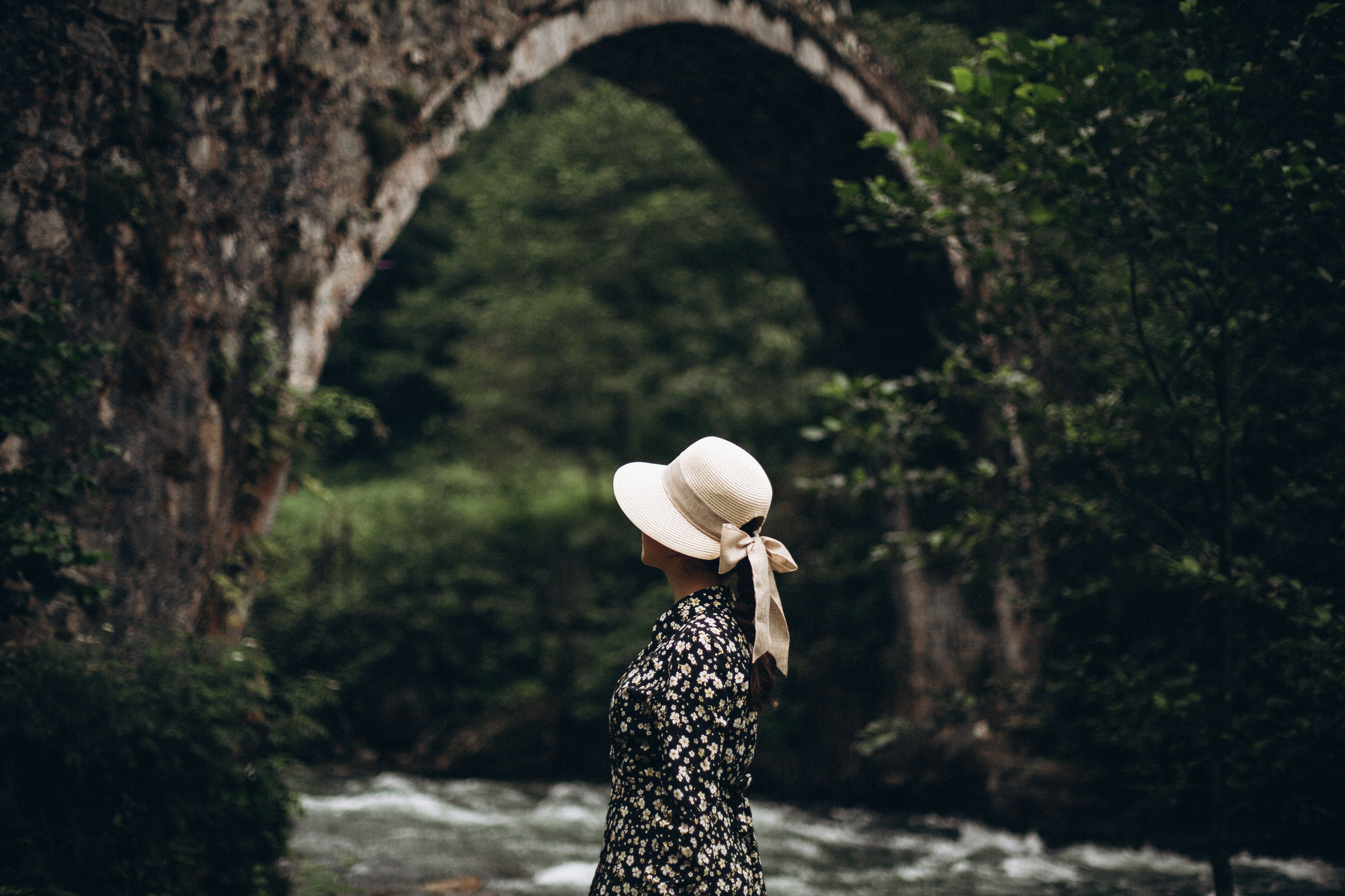 [ColoSach]-a-woman-in-a-floral-sundress-and-sunhat-gazes-at-a-stone-arch-bridge-in-a-lush-forest.