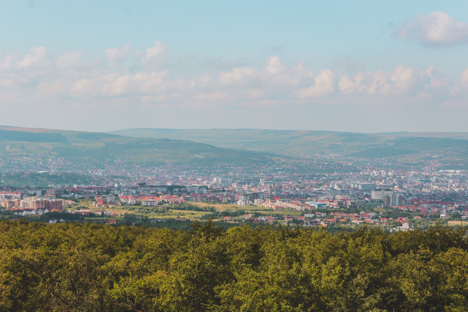 A panoramic cityscape view captured from a hilltop with dense greenery in the foreground.