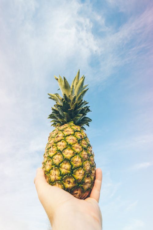 Free A human hand holding a ripe pineapple against a clear blue sky, symbolizing tropical freshness. Stock Photo