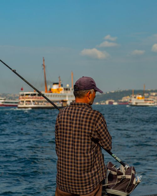 Free An elderly man enjoys fishing by the sea with a bustling cityscape and ships in the background. Stock Photo