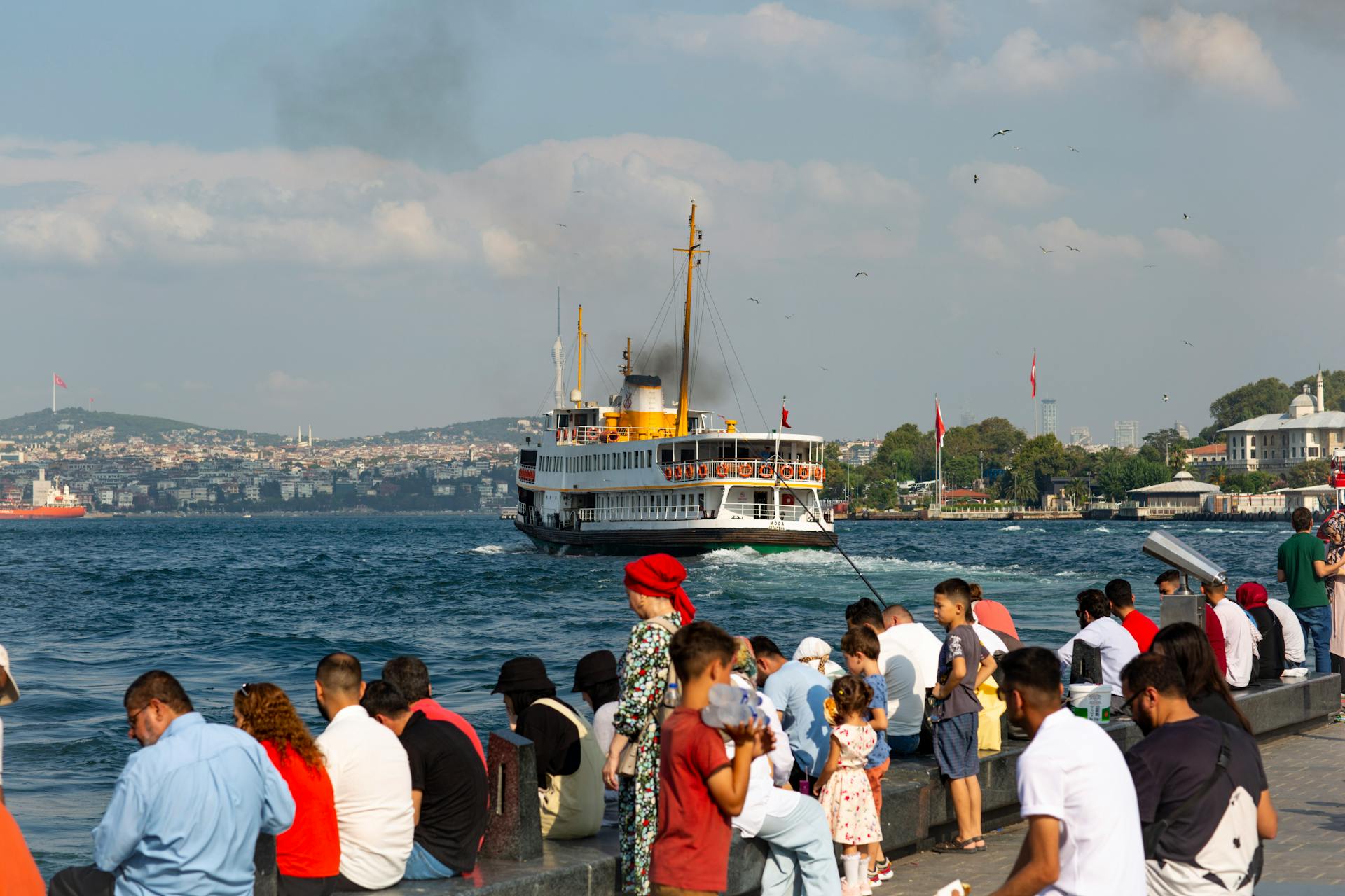 A bustling scene captures tourists and locals enjoying the Bosphorus Strait in Istanbul with a passenger ship approaching.