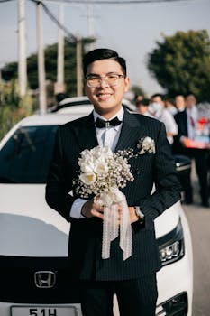 Smiling Asian groom in black suit holding bouquet, standing outdoors beside a white car, perfect for wedding themes.