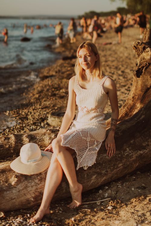Free Blonde woman in a white dress sitting on driftwood at a sunny beach, enjoying summer. Stock Photo