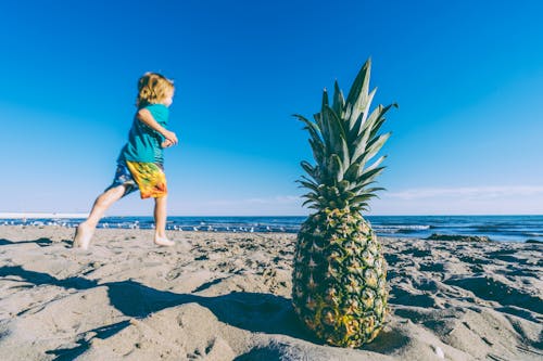 Child Running on Sand Near Bod of Water and Pineapple