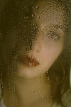 Close-up portrait of a woman viewed through a rain-covered window, capturing a serene and introspective mood.