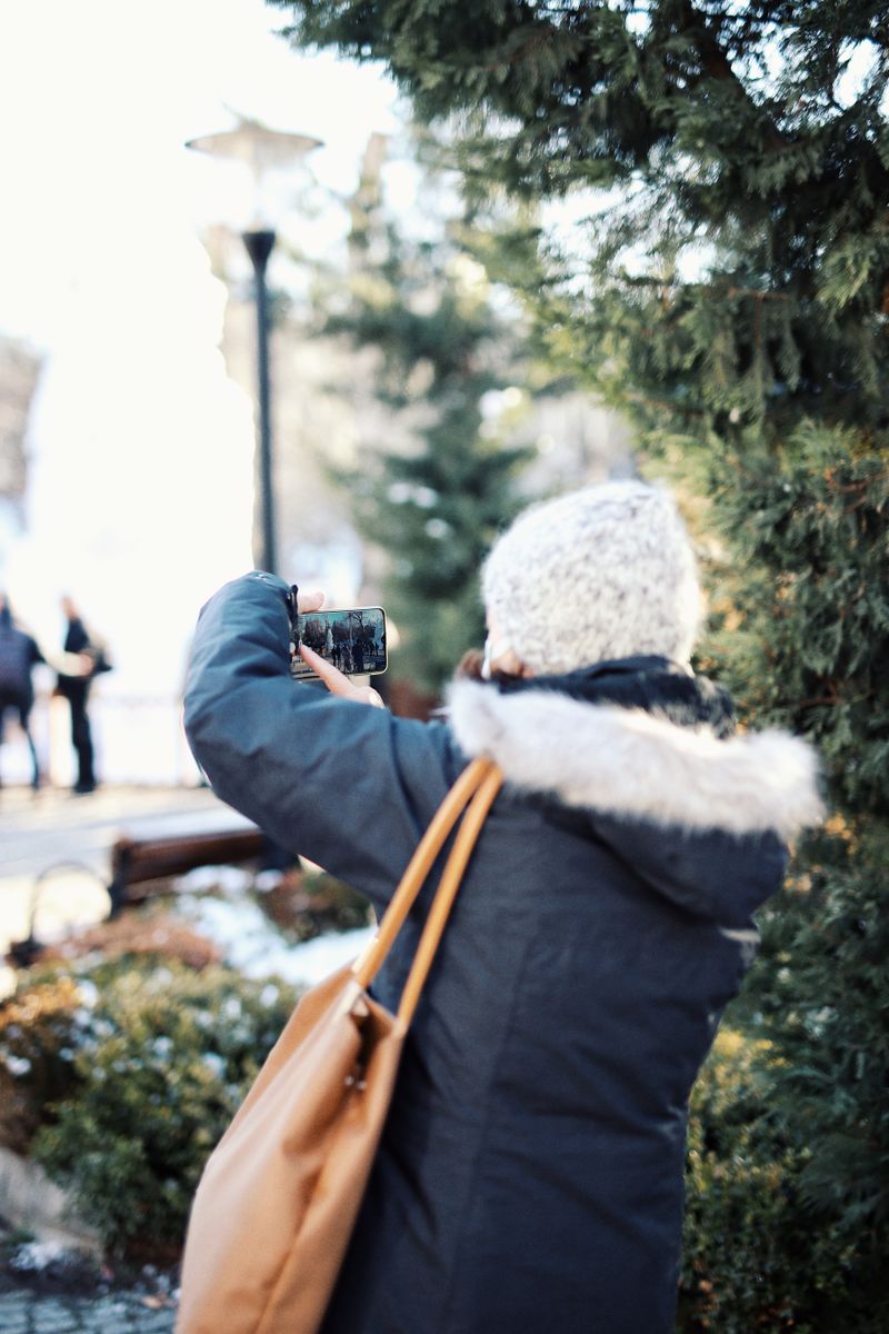 A woman in a winter coat photographing a park on her phone — photo by Rümeysa on Pexels