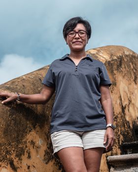 Confident woman wearing glasses and casual clothing, standing outdoors against a rock wall.