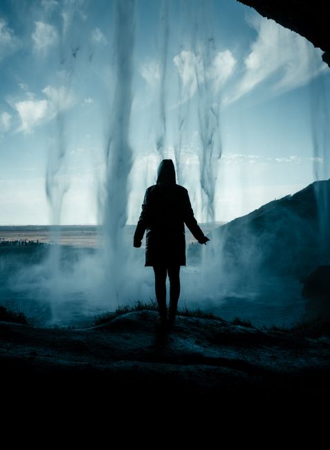 Person standing behind a cascading waterfall in Iceland with a stunning landscape view.