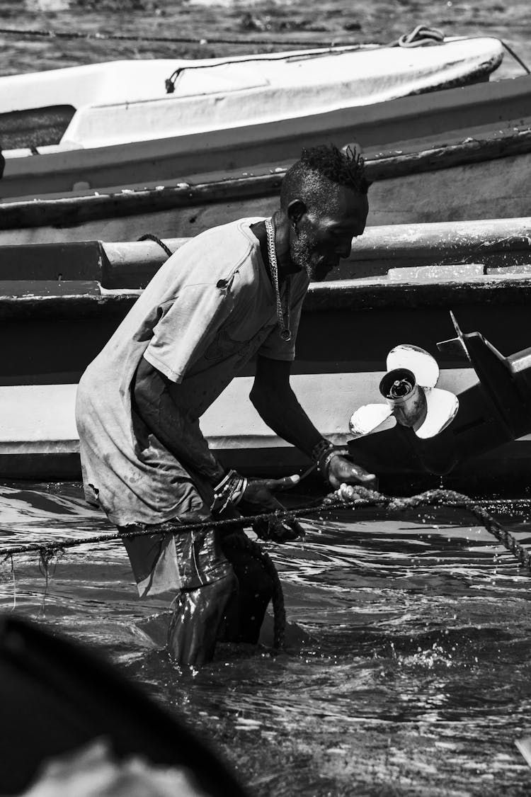 Grayscale Photo Of A Fisherman Near The Boats