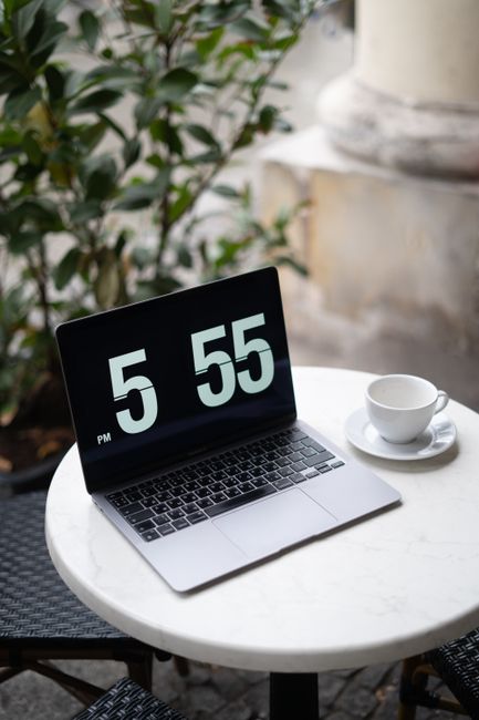 Laptop and coffee cup on a marble table at an outdoor café, displaying time 5:55 PM.