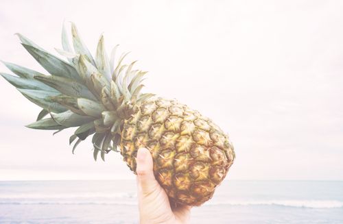 Free A hand holding a pineapple with a serene beach backdrop, perfect for travel and tropical themes. Stock Photo