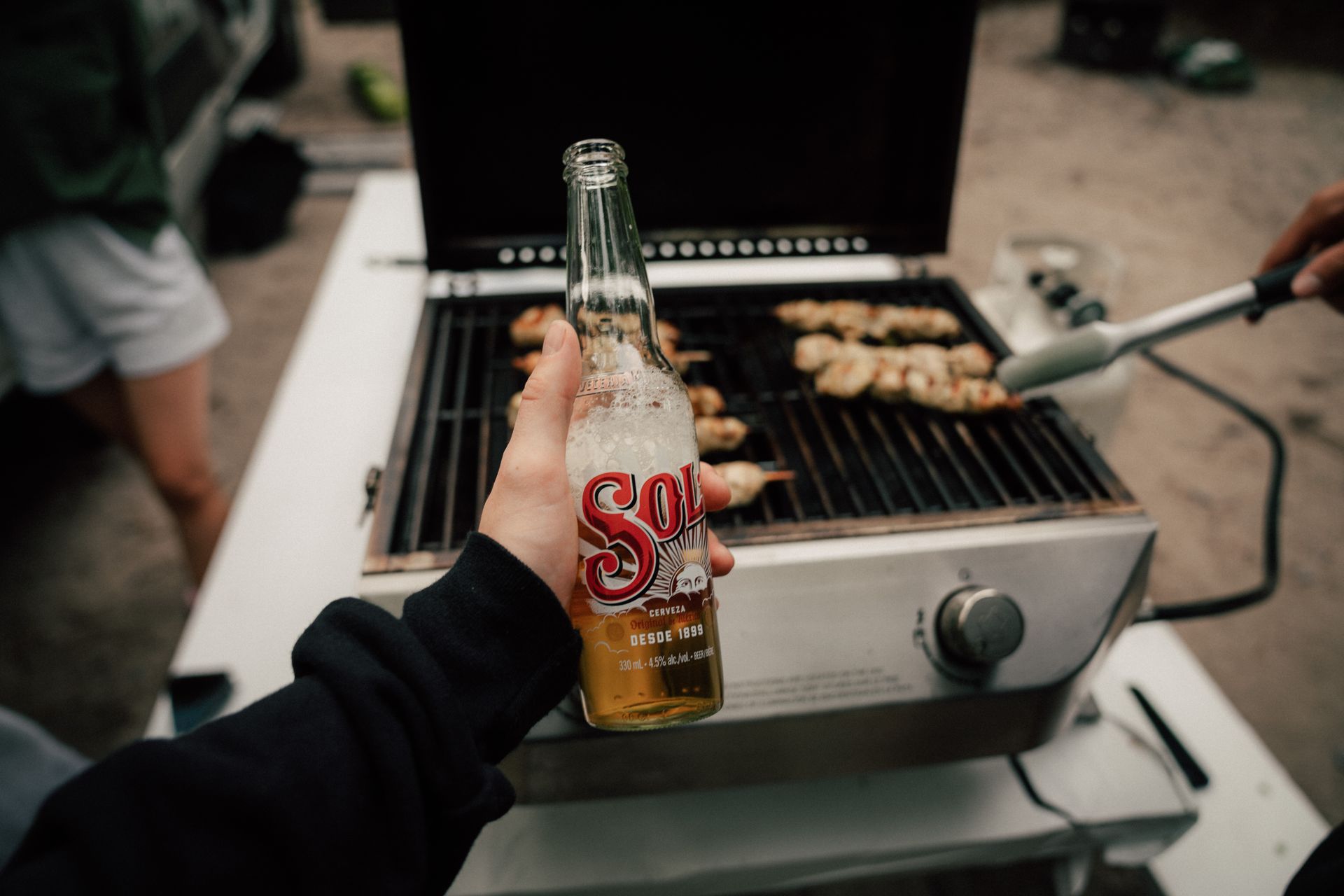 Refreshing Sol beer held in hand at a lively outdoor barbecue with grilled chicken skewers.