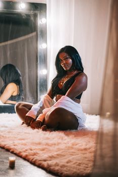 Elegant woman in black lingerie sitting on a furry rug, reflecting in a lit wall mirror.