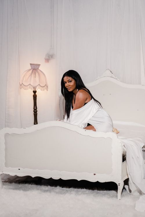 Free A graceful woman in a white dress poses elegantly on a bed in a softly lit room. Stock Photo