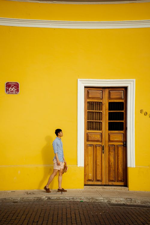 Free A young man walks past a colorful yellow building with a wooden door in Yucatán, Mexico. Captured outdoors. Stock Photo