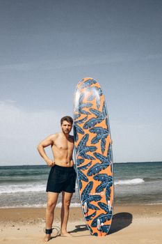 Caucasian man posing with a patterned surfboard on a sunny beach day.