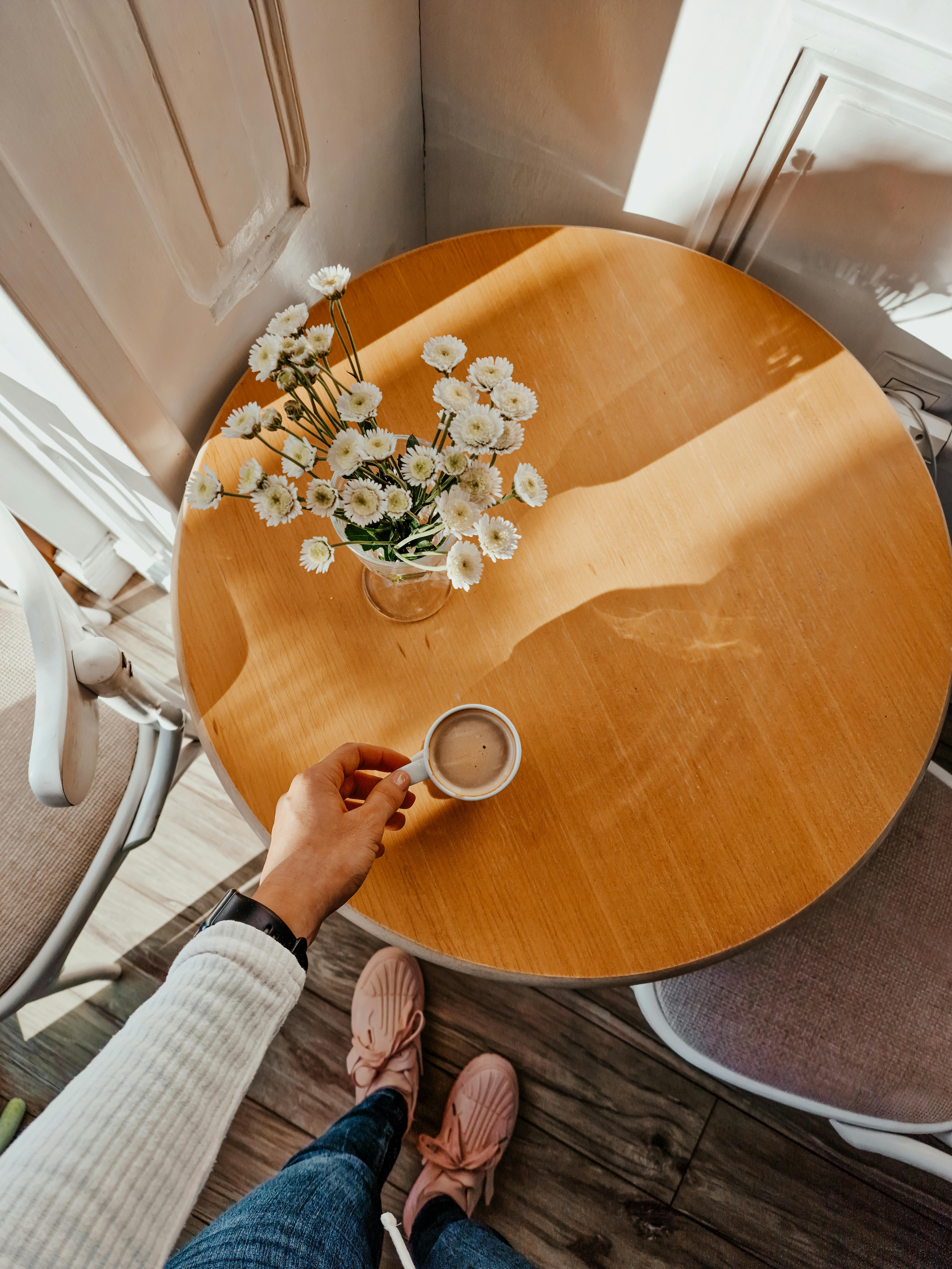 Free Top view of a hand holding coffee with daisies on a sunlit wooden table in Romania. Stock Photo