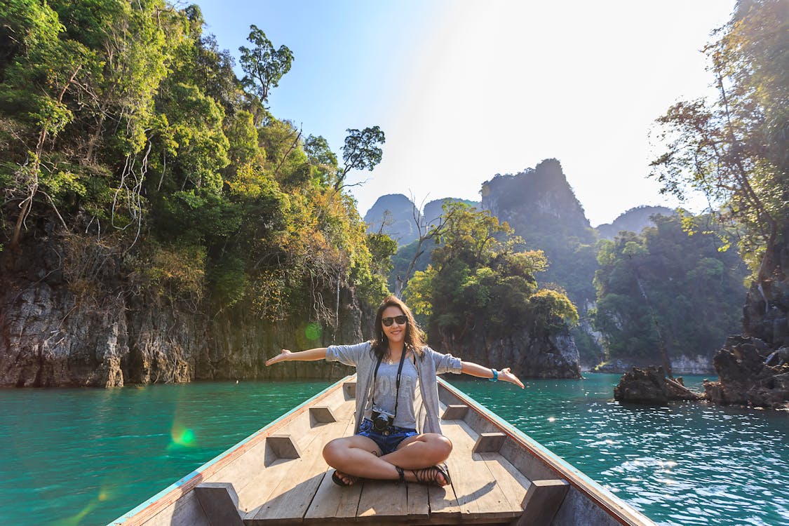 A young lady spreads her arms while sitting on a canoe against a backdrop of clear water and green mountains.