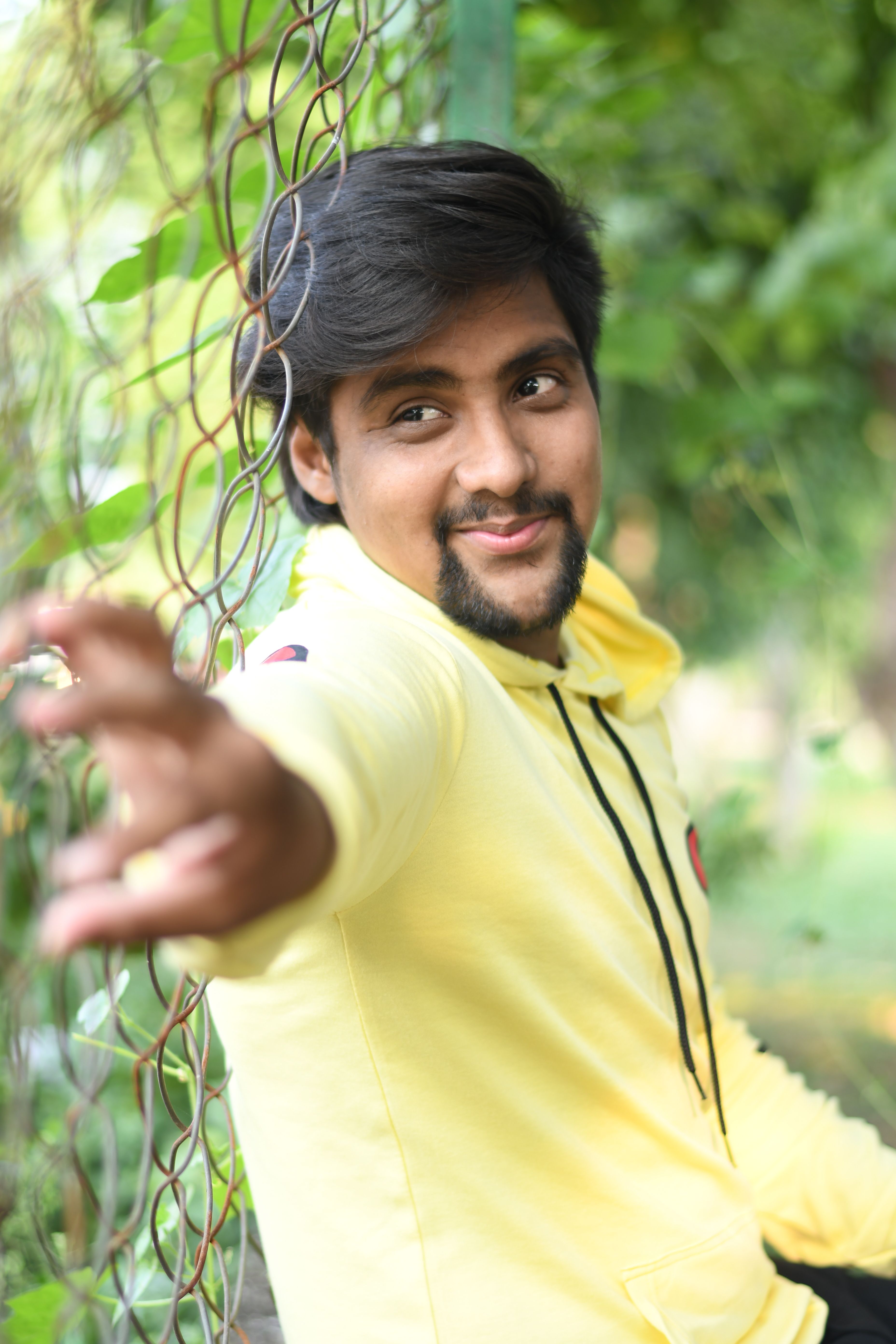 A cheerful young man reaching out with a smile, wearing a yellow hoodie near a fence outside.