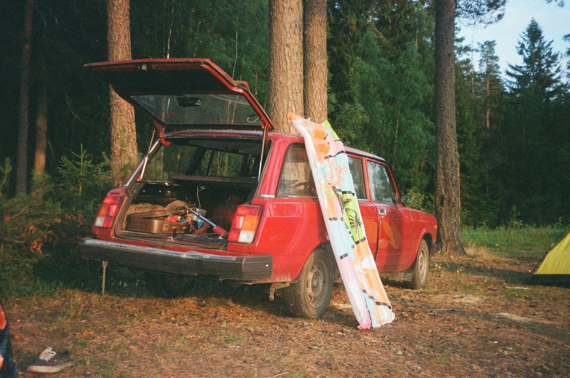 Vintage red car with open trunk in a forest, surrounded by camping gear and trees.
