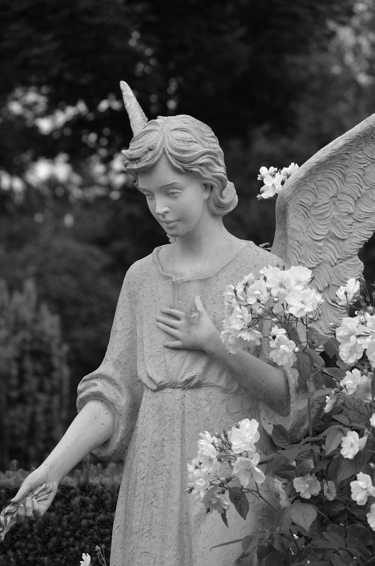 Grayscale Photograph Of An Angel Statue Near Flowers