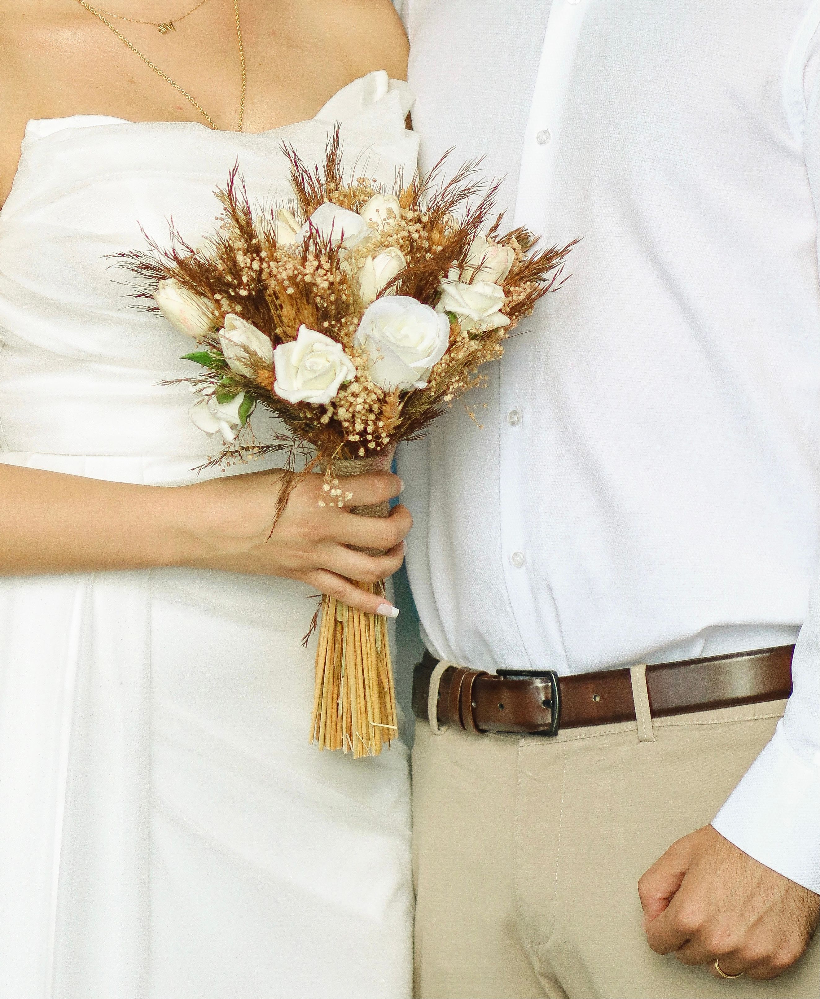 Close-up of a bride holding a rustic bridal bouquet alongside the groom in a white shirt.